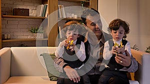 Portrait of middle-aged arabic father sitting on sofa with his twin sons playing with toy-robots in living room.