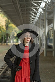 Portrait of middle-aged aristocratic woman in red dress, black coat and hat standing on platform of station waiting for train
