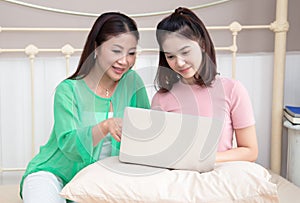 Portrait of middle aged Asian mother and teenage daughter using laptop computer, chat together while sitting on bed in bedroom at