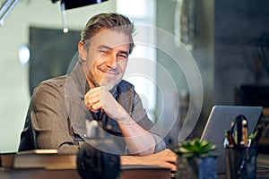 Portrait of middle aged bearded guy at home. Older handsome man smiling, looking at camera, working at home, using laptop computer