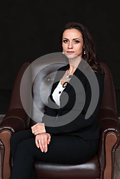 Portrait of middle aged business woman in formal black suit, sitting sideways on a armchair and looking seriously straight