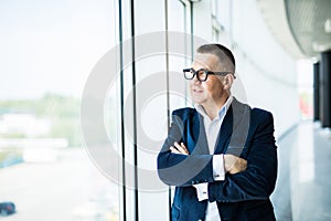 Portrait of a middle aged businessman in front of office window. Man is smiling and has his arms folded.