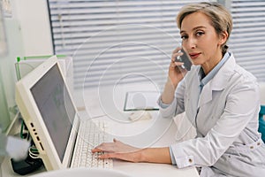 Portrait of middle-aged Caucasian female doctor in white coat talking on smartphone in hospital clinic sitting at desk