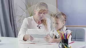 Portrait of middle aged caucasian woman and little schoolgirl sitting at the table with tablet. Tutor teaching