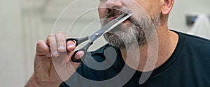 Portrait of Middle-aged handsome man cutting his beard with scissors. Selfcare during quarantine isolation