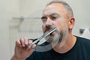 Portrait of Middle-aged handsome man cutting his beard with scissors. Selfcare during quarantine isolation