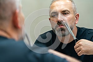 Portrait of Middle-aged handsome man cutting his beard with scissors. Selfcare during quarantine isolation