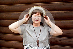 Portrait of a middle-aged happy woman bbw in a hat and linen dress near a log wall