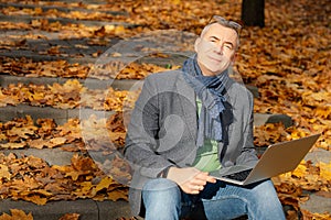 Portrait of middle-aged man sitting on concrete stairs covered with yellow fallen leaves in park forest, holding laptop.