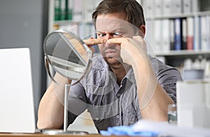 Portrait of middle-aged man sitting opposite small table mirror and squeezing acne problems