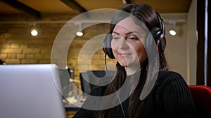Portrait of middle-aged overweight woman in headphones watching into laptop smilingly on office background. photo