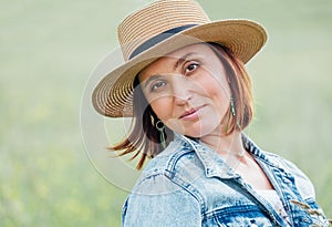 The portrait of a middle-aged smiling woman looking at the camera. She dressed in a jeans jacket, straw hat. Natural people beauty