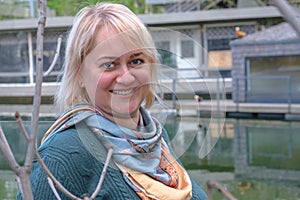 Portrait of a middle-aged woman with blond hair against the backdrop of a pond and a country house