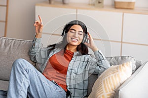 Portrait of millennial Arab woman in headphones listening to music with closed eyes, relaxing on couch at home