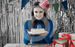 Portrait of a millennial girl sitting at a table with a birthday cake in her hands and a blown out candle. A young lady