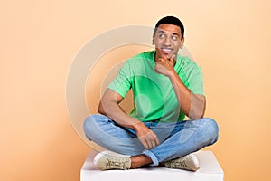 Portrait of minded man wear stylish t-shirt jeans sit on white cube thoughtfully look empty space isolated on beige