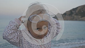 Portrait of mischievous caucasian blonde baby girl wearing glasses for swimming and looking at the sea.