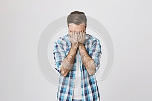 Portrait of miserable and sad male adult, covering face with hands, expressing panic and depression over gray background