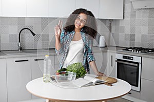 Portrait of mixed-race curly teen girl standing in the kitchen in front of a table with vegetables and a book for