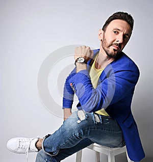 Portrait of modern adult unshaved man in torn jeans, yellow t-shirt and blue jacket sitting on chair with legs crossed