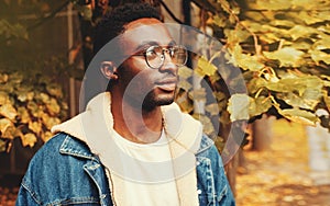Portrait of modern african man thinking and looking away wearing eyeglasses in autumn city park on yellow leaves background