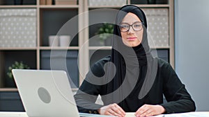 Portrait of modern ethnic Muslim young woman posing at home office workplace sitting at table