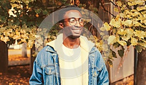 Portrait of modern happy smiling african man looking away wearing eyeglasses in autumn city park on yellow leaves background