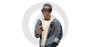 Portrait of modern young african man with smartphone wearing jean jacket, sunglasses and backpack isolated on white background