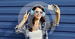 Portrait of modern young woman taking selfie picture by smartphone in wireless headphones listening to music over blue