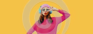 Portrait of modern young woman in wireless headphones listening to music wearing knitted sweater, pink hat on yellow background