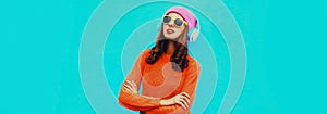 Portrait of modern young woman in wireless headphones listening to music wearing red knitted sweater, pink hat on blue background