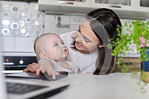 Portrait of mom and baby son sitting at home in kitchen looking in laptop screen