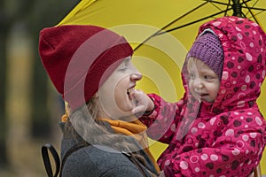 Portrait of mom and baby under yellow umbrella in park. Mom and little daughter fooling around