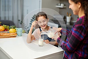 Portrait of a mom pours her cute laughing daughter milk in glass for breakfast