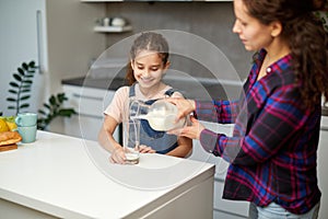 Portrait of a mom pours her cute smiling daughter milk in glass for breakfast.