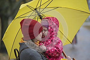 Portrait of mom who hugs her little daughter holding her in her arms under yellow umbrella in a yellow autumn park