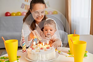 Portrait of mother and baby with birthday cake sitting at table and looking at delicious dessert, celebrating first kid`s birthda