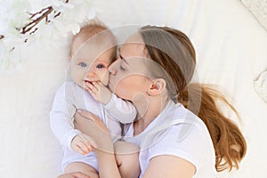 Portrait of a mother with a baby, a mother kisses a child and gently hugs him on a white bed at home, maternal love and care