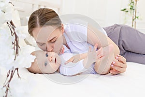 Portrait of a mother with a baby, a mother kisses a child and gently hugs him on a white bed at home, maternal love and care