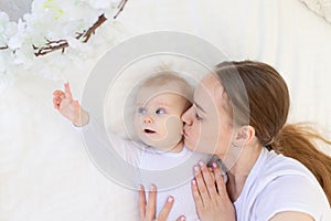 Portrait of a mother with a baby, a mother kisses a child and gently hugs him on a white bed at home, maternal love and care