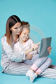 Portrait of mother and daughter in pajamas with tablet sitting on the floor isolated over the blue studio.