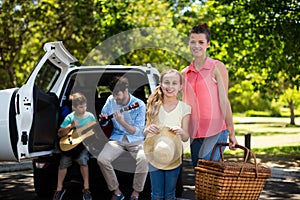 Portrait of mother and daughter standing with picnic basket while father and son playing guitar in b