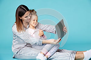 Portrait of mother and daughter with tablet sitting on the floor isolated over the blue studio.