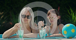 Portrait of a mother with her daughter, relaxing in the pool. Cooling drinks are next to them. Looking at the camera