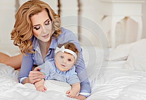 Portrait of mother and little baby girls with blue eyes in a striped blue dress lying on white bed. Mom tenderly looking at her d