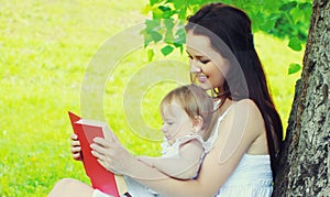 Portrait mother and little girl baby reading a book together on the grass in a summer park