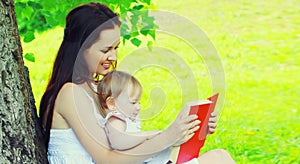 Portrait of mother and little girl baby reading book together on the grass under tree in summer park