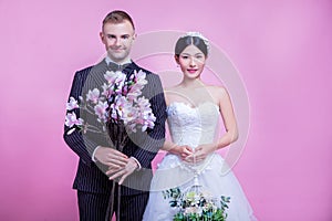 Portrait of multi-ethnic wedding couple holding flowers while standing against pink background