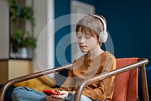 Portrait of music lover teen boy listening to music in headphones sitting on armchair at home.