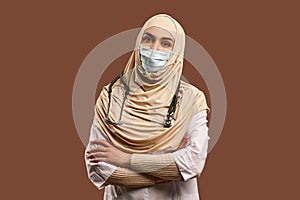 Portrait of Muslim doctor woman wearing white robe and protective mask, arms crossed, standing near brown background. The concept